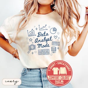 May include: Ivory-colored t-shirt with the text "Data Analyst Mode" in navy blue, accompanied by graphic elements like charts, a pie chart, and a database icon. The shirt also features a Comfort Colors logo.