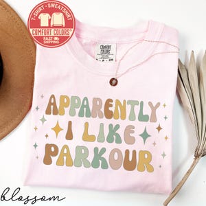 May include: A light pink t-shirt with the text "APPARENTLY I LIKE PARKOUR" in retro-style lettering. The shirt has a crew neck and short sleeves. A gold necklace with a pendant is visible. A red and white sticker is in the upper left corner.