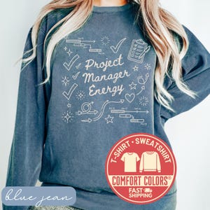 May include: Blue jean colored sweatshirt with the words "Project Manager Energy" in white script. The design includes checkmarks, arrows, and a clipboard graphic. A red circular logo at the bottom reads "T-Shirt Sweatshirt Comfort Colors Fast Shipping."