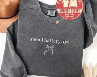 May include: A dark gray Comfort Colors sweatshirt with the text "social battery: 0%" and a bow graphic. The sweatshirt has a crew neck and long sleeves. A red and white circular graphic reads "T-Shirt Sweatshirt" and "Comfort Colors".