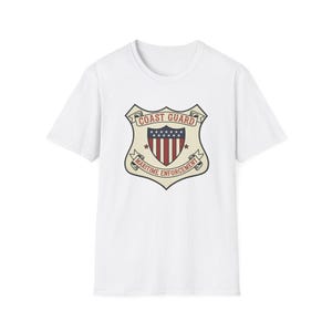 May include: White t-shirt featuring a Coast Guard emblem. The emblem is shield-shaped with a red and white striped center, a blue field of stars, and the words "Coast Guard" and "Maritime Enforcement".