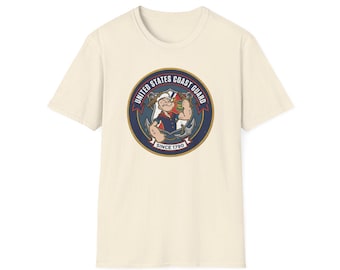 May include: Cream-colored t-shirt featuring a circular graphic of Popeye in a U.S. Coast Guard uniform. The graphic includes the text "UNITED STATES COAST GUARD" and "SINCE 1790" in a navy blue and red design. The shirt has short sleeves.