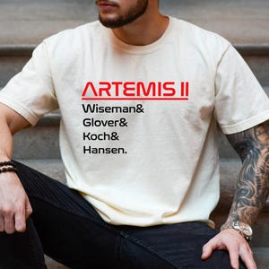 May include: Cream-colored t-shirt with the text "ARTEMIS II" in red, followed by the names Wiseman, Glover, Koch, and Hansen in black. The shirt is worn by a person wearing black jeans and a watch.