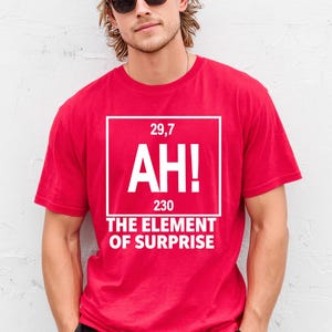 Puede incluir: Una camiseta roja con el texto "AH! THE ELEMENT OF SURPRISE" en un cuadrado blanco. La camiseta también presenta los números "29,7" y "230". La persona de la imagen lleva una gorra negra y gafas de sol.