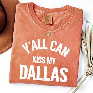 Può includere: Una t-shirt a maniche corte color terracotta con scollo rotondo e testo stampato in bianco che recita Y'ALL CAN KISS MY DALLAS. La maglietta presenta un'etichetta del marchio Comfort Colors all'interno del collo e una taglia S visibile.