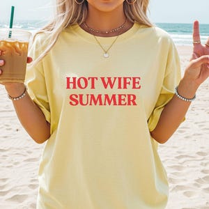 May include: A pale yellow t-shirt with the red text "HOT WIFE SUMMER". The image shows the shirt on a beach with the sea in the background. The person is holding a drink and making a peace sign.