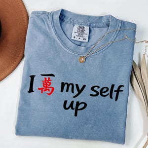 May include: A blue Comfort Colors t-shirt with the phrase "I 萬 my self up" printed in black and red. A gold necklace with a pendant is draped over the shirt. A brown hat and dried leaves are in the background.