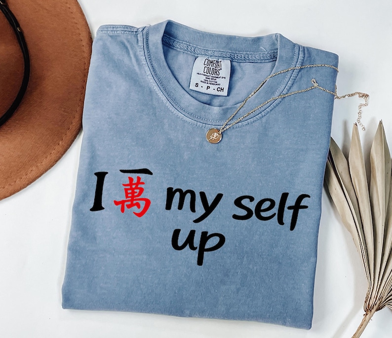 May include: A blue Comfort Colors t-shirt with the phrase "I 萬 my self up" printed in black and red. A gold necklace with a pendant is draped over the shirt. A brown hat and dried leaves are in the background.