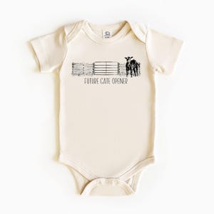 Pode incluir: Body de bebé creme com um gráfico preto de uma cerca, duas vacas e o texto "FUTURE GATE OPENER". O body tem mangas curtas e fecho de pressão na parte inferior.