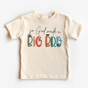 May include: Cream-colored toddler t-shirt with the phrase "so God made a BIG BRO" in black script. The words "BIG BRO" are in colorful block letters with farm-themed illustrations. A tractor, cow, and truck are included.