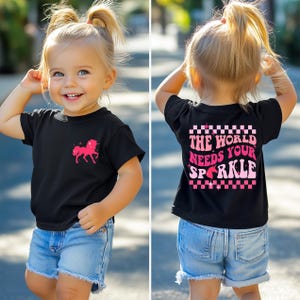 Puede incluir: Camiseta negra para niño con un diseño de unicornio rosa y la frase "THE WORLD NEEDS YOUR SPARKLE". La camiseta se combina con pantalones cortos de mezclilla. El niño sonríe.