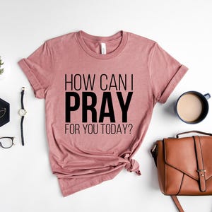 May include: A dusty rose t-shirt with the text "HOW CAN I PRAY FOR YOU TODAY?" is displayed on a white surface, alongside a brown purse, a cup of coffee, and other accessories. The shirt is knotted at the bottom.