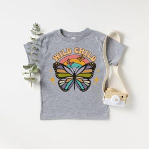 May include: A gray t-shirt with the words "WILD CHILD" above a colorful butterfly graphic. The butterfly has black outlines and wings in shades of blue, green, yellow, and purple. A rainbow and small butterflies are above the butterfly.