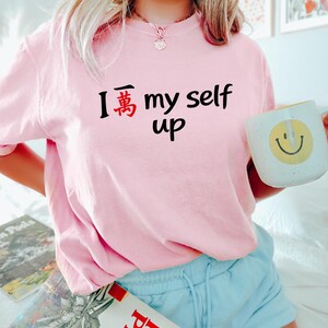 May include: A light pink t-shirt with the text "I 萬 my self up" in black and red. A white mug with a yellow smiley face is held in the right hand. The person is wearing light blue shorts.