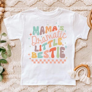May include: White t-shirt with the text "Mama's Dramatic Little Bestie" in colorful, retro-style lettering. The design includes stars and a checkered border. A cute and playful design for kids.