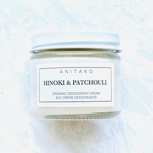 Op de afbeelding: Een kleine, heldere glazen pot met een zilveren deksel, met biologische deodorantcrème. Het etiket luidt "ANITAKO HINOKI & PATCHOULI" met extra tekst in het Engels en Frans. De pot staat op een wit en blauw getextureerd oppervlak.