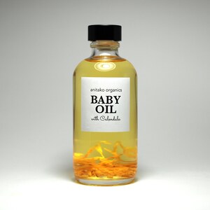 Pu&ograve; includere: Una bottiglia di vetro trasparente di olio per bambini con un tappo nero. L'etichetta recita "anitako organics BABY OIL with Calendula".