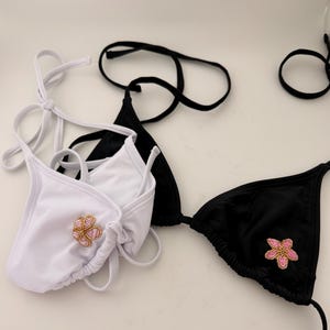 May include: White and black bikini tops with floral embellishments. The white bikini top has a pink and gold flower design. The black bikini top has a pink flower design. Both tops have tie closures.