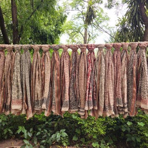 May include: A display of patterned scarves in shades of brown, beige, and red, suspended from a rod. The scarves showcase detailed designs, including floral and geometric patterns. The backdrop is a vibrant green with trees and foliage.