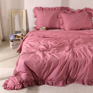 Op de afbeelding: Een roze beddengoedset met ruches. De set bevat een dekbedovertrek en twee kussenslopen. Het dekbedovertrek heeft houten knopen. Een klein wit kopje staat op het bed. Een spiegel en een mandje staan op de achtergrond.