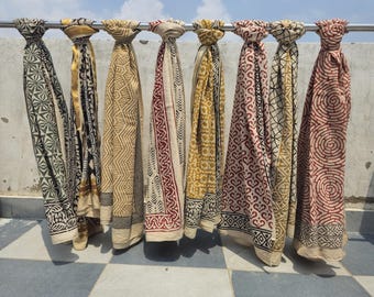 May include: A display of patterned scarves in shades of green, yellow, and red, suspended from a metal rail. The scarves showcase geometric and floral patterns, with a blend of light and dark hues. The backdrop is a clear, blue sky.