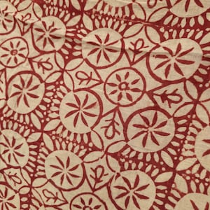 May include: Close-up of a cream-colored fabric with a repeating pattern of red floral and circular designs. The pattern includes stylized flowers, leaves, and dotted circles, creating a textured, artistic look. The fabric appears to be lightweight.