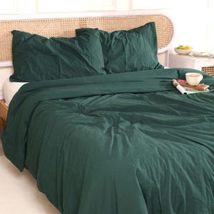 May include: A dark green bed set, including a duvet cover and two pillowcases, is displayed on a bed. The bedding has a slightly wrinkled texture. A white nightstand with a cup and a magazine is visible.