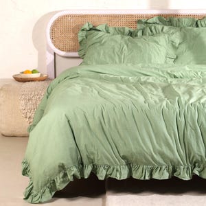 May include: A sage green bed set with ruffled edges. The set includes a duvet cover and pillowcases. The bed is made up with a light-colored headboard and a small wooden tray with fruit.