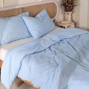 May include: A light blue bed set, including two pillows and a duvet, arranged on a bed. The bedding has a slightly wrinkled texture. The headboard is made of woven wicker. The overall theme is bedroom decor.