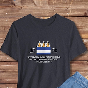 May include: Dark navy blue t-shirt with a graphic of three beer bottles in a box, and the quote "WE'RE FAMILY - WE'RE GONNA BE DOING LOTS OF DUMB STUFF TOGETHER!" by Tommy Callahan. Light blue jeans are folded next to the shirt.