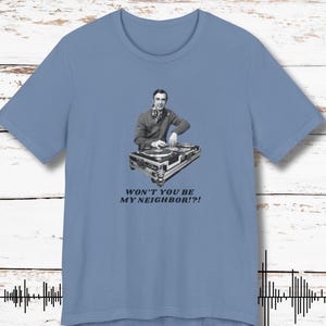 May include: A light blue t-shirt featuring a black and white image of a person with headphones at a turntable. The text "WON'T YOU BE MY NEIGHBOR!?" is printed below the image. The shirt is made of a soft material.