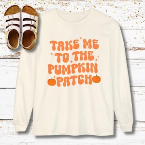 May include: A long-sleeved cream-coloured shirt featuring the orange text "TAKE ME TO THE PUMPKIN PATCH" with small star accents and two orange pumpkin illustrations. Beside the shirt are two white sandals with three adjustable buckle straps.