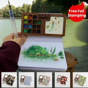 May include: A watercolour painting kit with a brown palette containing various colours, a water brush, and a sketchbook with a landscape painting. The kit is held open, displaying the art supplies and a finished painting. The image also shows a red circle with the text "Free Foil Stamping."
