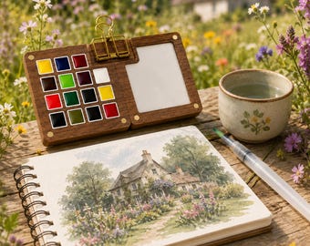 May include: A wooden watercolor palette box with sixteen small square paint pans in various colors, a gold metal binder clip, a white mixing area, a spiral-bound sketchbook featuring a watercolor painting of a cottage, and a water brush pen.