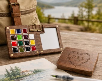 May include: A dark wood watercolor paint set featuring twelve square paint pans in various colors, a white mixing area, and a metal binder clip. The closed wooden lid is engraved with a heart, the letters D & L, and the date 06.23.24.