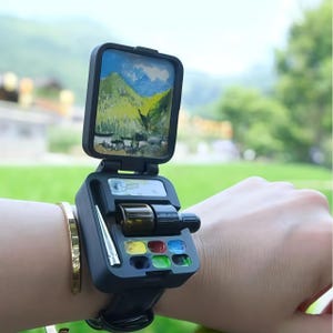 Może przedstawiać: Czarny zegarek na rękę z miniaturowym obrazem krajobrazu na górze. Tarcza zegarka otwiera się, ukazując małe butelki i pojemniki na farby w kolorach czerwonym, żółtym, niebieskim i zielonym. Dołączone jest również srebrne narzędzie.