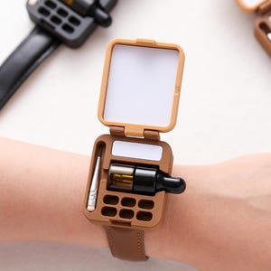 Może przedstawiać: Brązowa bransoletka z prostokątnym etui, które otwiera się, odsłaniając lustro i przegródki. Etui zawiera małą fiolkę i czarną butelkę. Bransoletka jest wykonana ze skóry w kolorze brązowym.