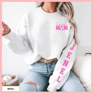 May include: White sweatshirt with the text "Soccer Mom" in pink on the chest, and the name "JENEL" in pink down the left sleeve. The sweatshirt is paired with blue jeans.