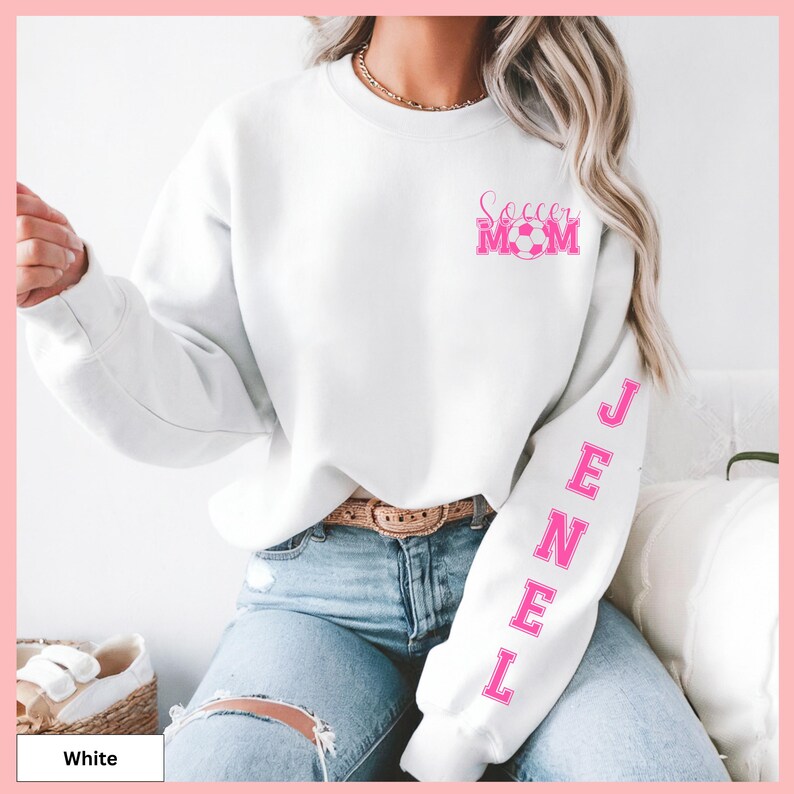 May include: White sweatshirt with the text "Soccer Mom" in pink on the chest, and the name "JENEL" in pink down the left sleeve. The sweatshirt is paired with blue jeans.