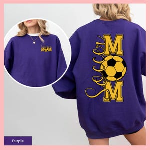 May include: A purple sweatshirt with "Soccer Mom" printed on the back in yellow and black. The design includes a soccer ball and the words "Soccer Mom". The front of the sweatshirt has the words "Soccer Mom" in a smaller design.