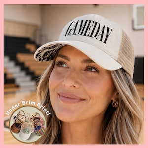May include: A cream-colored baseball cap with a mesh back and the word GAMEDAY printed in black on the front. The underside of the brim features a black and white animal print pattern. A small circular inset shows various other cap designs.
