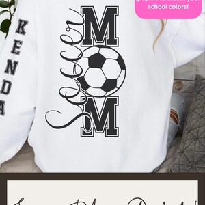 May include: White sweatshirt with a black soccer ball graphic and the words "Soccer Mom" in a stylized font. The word "MOM" is in bold block letters. The sleeve has the name "KENDA" in block letters. The text "Soccer Mom Outfits!" is at the bottom.