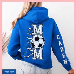 May include: Royal blue hoodie with the text "Soccer Mom" and a soccer ball graphic on the back. The name "Caden" is printed vertically on the right sleeve. The hoodie is a solid royal blue color.