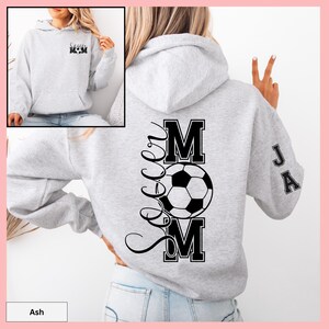 May include: Light gray hoodie with a front pocket and a hood. The back features a large black graphic that says "Soccer Mom" with a soccer ball in the center. The sleeve has the initials "JA" in black. The front of the hoodie has the words "Soccer Mom" in black.