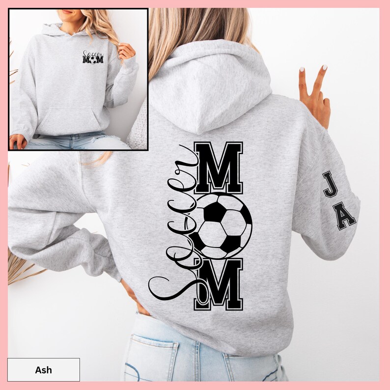 May include: Light gray hoodie with a front pocket and a hood. The back features a large black graphic that says "Soccer Mom" with a soccer ball in the center. The sleeve has the initials "JA" in black. The front of the hoodie has the words "Soccer Mom" in black.