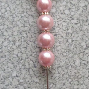 Puede incluir: Un alfiler blanco en forma de corazón con un diseño floral y detalles de perlas rosas. El alfiler tiene un tallo de metal plateado.