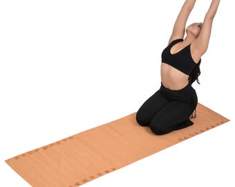 Op de afbeelding: Een persoon in een zwarte sport-bh en zwarte broek knielt op een rechthoekige, lichtbruine yogamat. De armen zijn boven het hoofd uitgestrekt. De mat heeft een textuur en decoratieve elementen langs de randen.