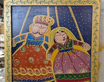 May include: Hand-painted wooden wall art featuring a couple in traditional Indian attire. The artwork has a gold border with a blue background and intricate details. The figures are dressed in vibrant colors, including orange, blue, and purple.