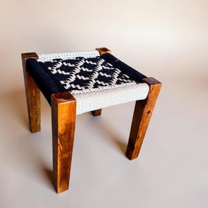 May include: A small square stool with four straight wooden legs in a medium brown finish. The seat features a woven cord pattern in contrasting black and off-white colors, creating a geometric diamond design across the top surface of the stool.
