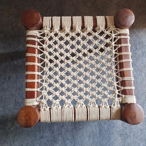 May include: A small, handcrafted wooden stool with a woven rope seat. The frame is made of dark brown wood, with rounded legs at each corner. The seat is a cream-colored, open weave design.